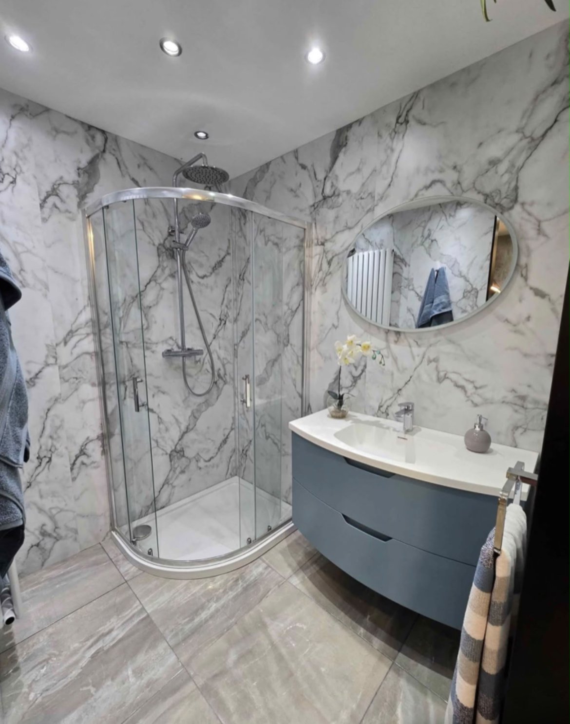 Modern marble bathroom with curved glass shower enclosure, curved sink vanity with dark base, round mirror, and recessed ceiling lights